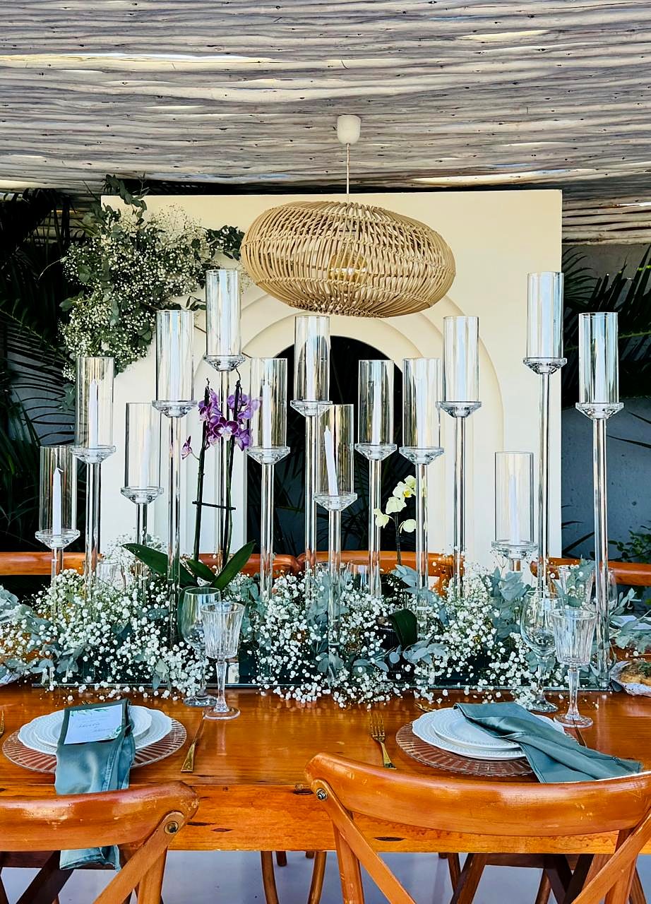 Castiçais de vidro de várias alturas com gipsófila, orquídeas e candeeiro de vime sobre mesa de madeira
