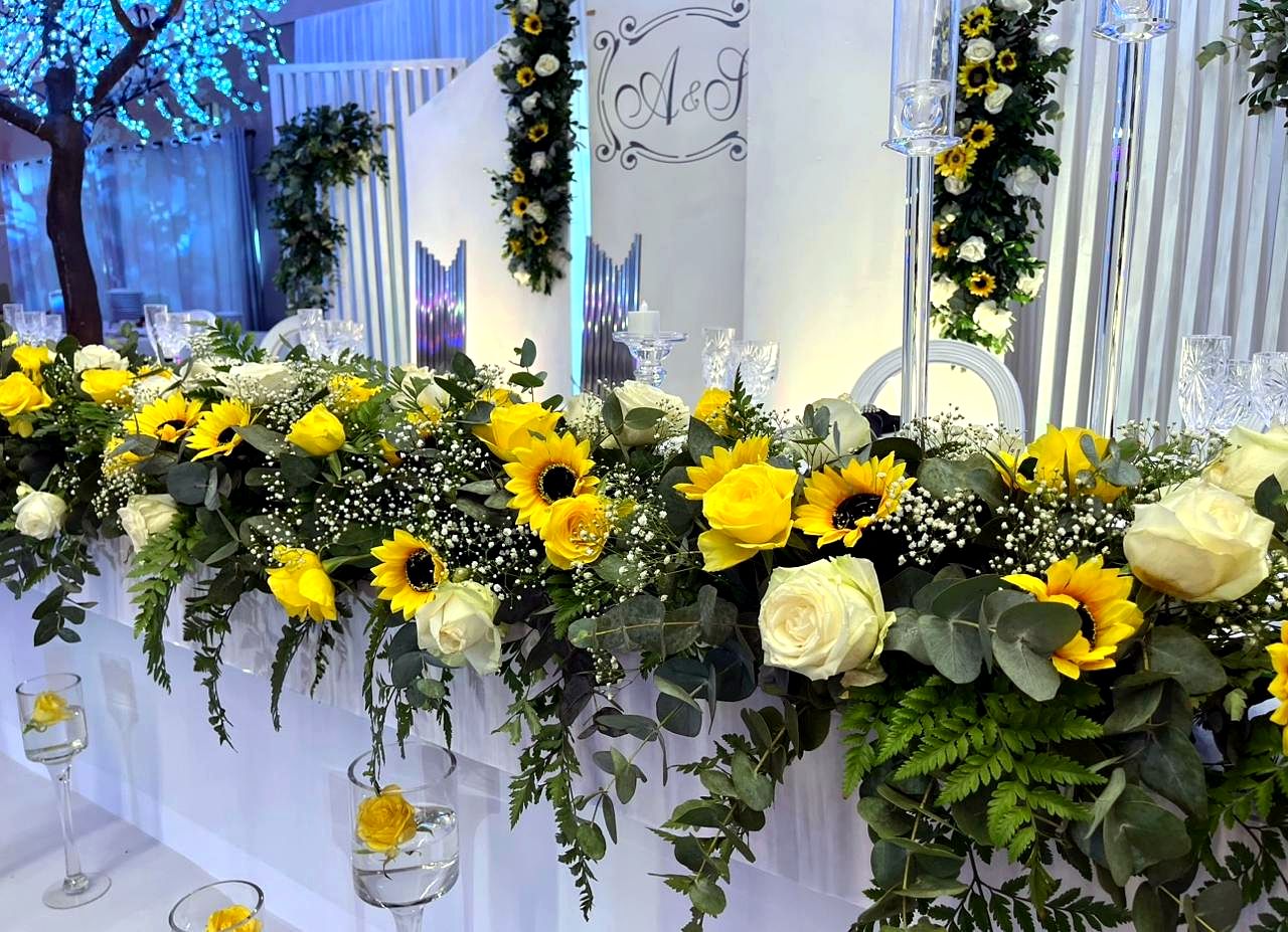 Mesa principal com girassóis e rosas brancas, árvore com luzes azuis ao fundo